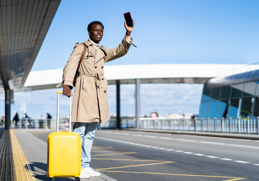 A young man with a phone in his hand and yellow luggage hails a taxi at the airport.